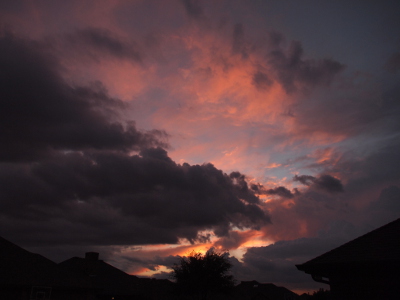 [Above the rooftops are clouds at several different layers from the foreground to the background. The foreground clouds are completely violet from a recent storm. Behind them are red/orange clouds in the lower portion and pink clouds in the upper portion as the setting sun lights them. Portions of blue sky are visible in the background.]